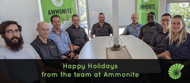 Ammonite team sitting in the boardroom. From left to right: Kyal, Jim, Rick, Scott, Mike,  Tim, Micah, Ben and Kylie. The Ammonite signs are in the background and the text below reads Happy Holidays from the team at Ammonite with the Ammonite logo on the right of the text